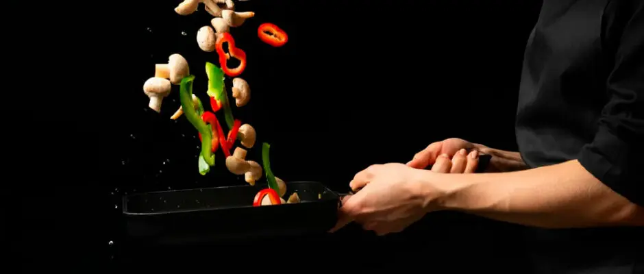 Las manos de un chef vestido de negro, mueven un sartén mientras saltean Champiñones Setas de Cuivá y pimentones. Realiza deliciosas Recetas