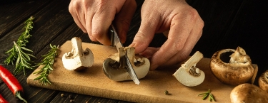 Sobre una tabla de madera, las manos de un chef sostienen un cuchillo que corta champiñones de Setas de Cuivá